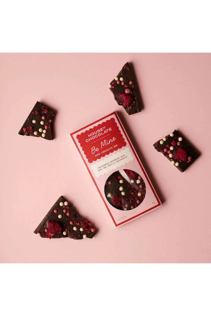 House of Chocolate Raspberry and Crispearl Dark Chocolate Bar.  Bar laying flat wrapped in a Pink and Red Sleeve reading Be Mine with another bar broken up laying around the flat laid packaged bar.