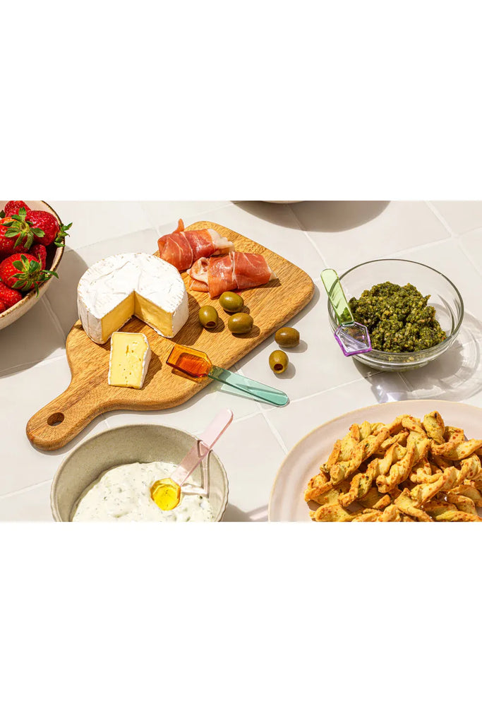 Sucker Multi-coloured Glass spoons shown on a chauteria board. Assorted food items including cheese, olives, and bread on a wooden board with a bowl of pesto and a plate of fried snacks.