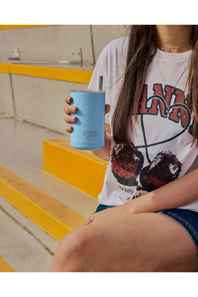 Person holding a blue tumbler with a Frank Green logo on it wearing a a white shirt.