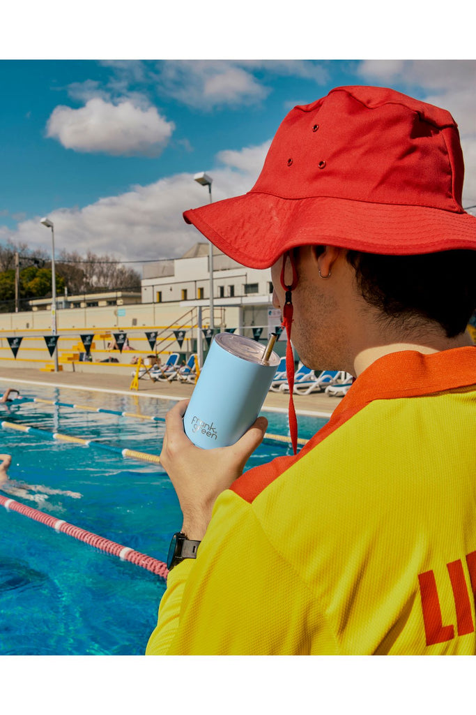Person in a yellow life vest holding a blue reusable cup with a Frank Green logo on it by a pool