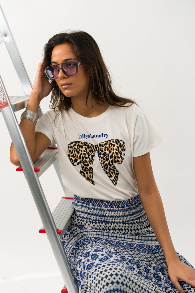 Woman wearing a white t-shirt with a leopard print bow design and patterned pants, sitting on a ladder. Lollys Laundry Roma Tee White.