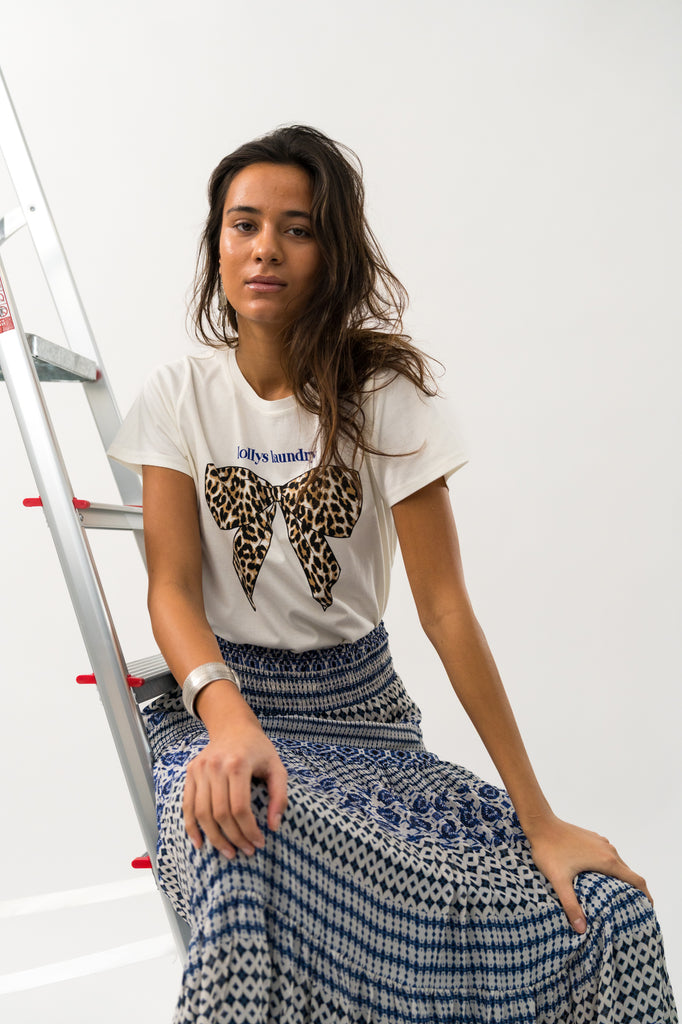 Woman wearing a white t-shirt with a leopard print bow design and patterned pants, sitting on a ladder. Lollys Laundry Roma Tee White.