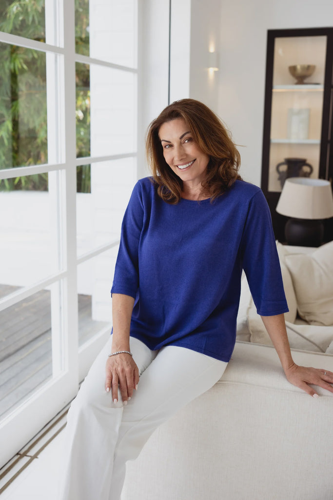 Woman in a blue top and white pants sitting on a couch in a modern living room. Alashan Everyday Tee in Royal.