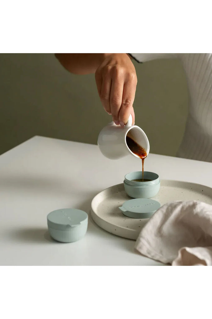 Person pouring a salad dressing from a white jug into one of Stylewares Eucalyptus Green mini containers on a light surface.