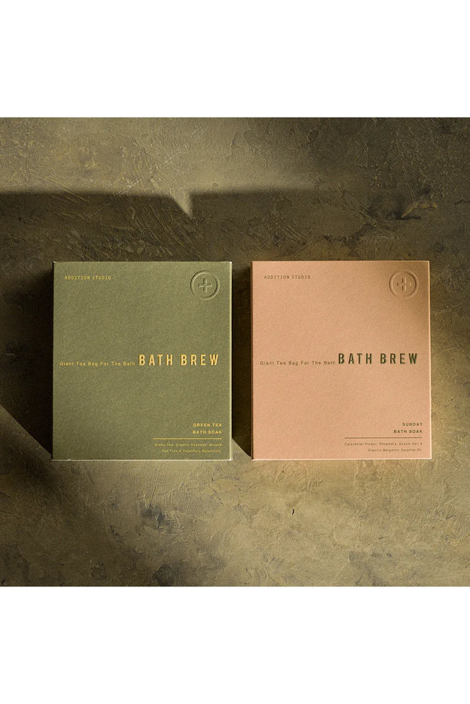 Addition Studio Giant Tea Bag For The Bath, Bath Brew. Two Bath Brew packages, one green and one pink, on a textured surface.