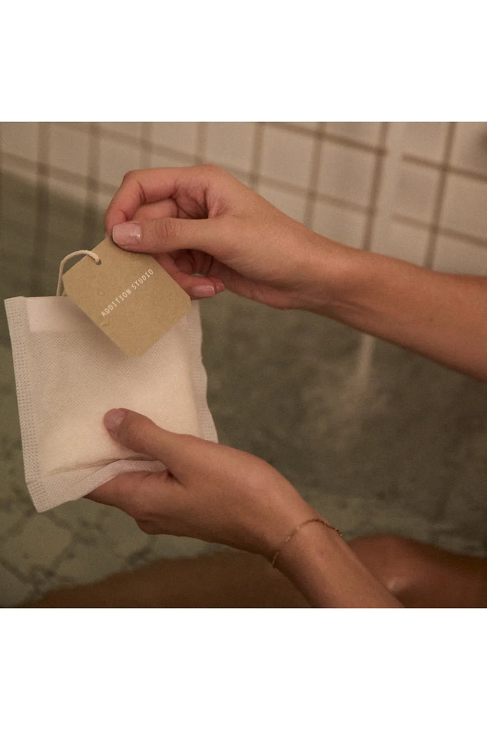 Addition Studio Giant Bath Brew Tea Bag: a person holding a woven tea bag with a brown tag against a tiled wall background in the bath.