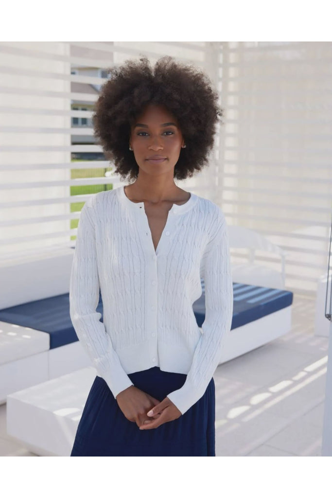 Woman wearing a white ribbed sweater and dark blue skirt standing in a bright outdoor setting. Alashan April Cable Cardigan in White.