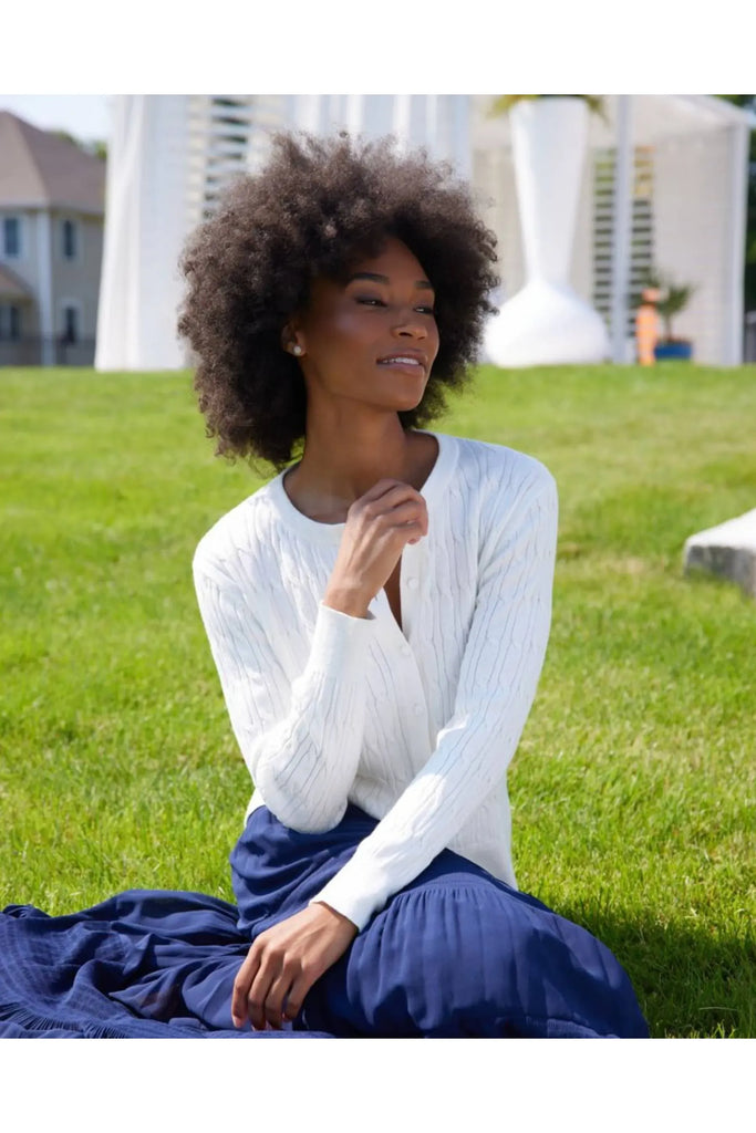 Woman wearing a white ribbed sweater and dark blue skirt standing in a bright outdoor setting. Alashan April Cable Cardigan in White.