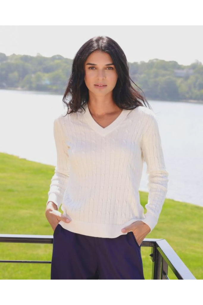Woman wearing a white sweater standing by a lake with greenery in the background. Alashan White April v Neck Cable Jumper