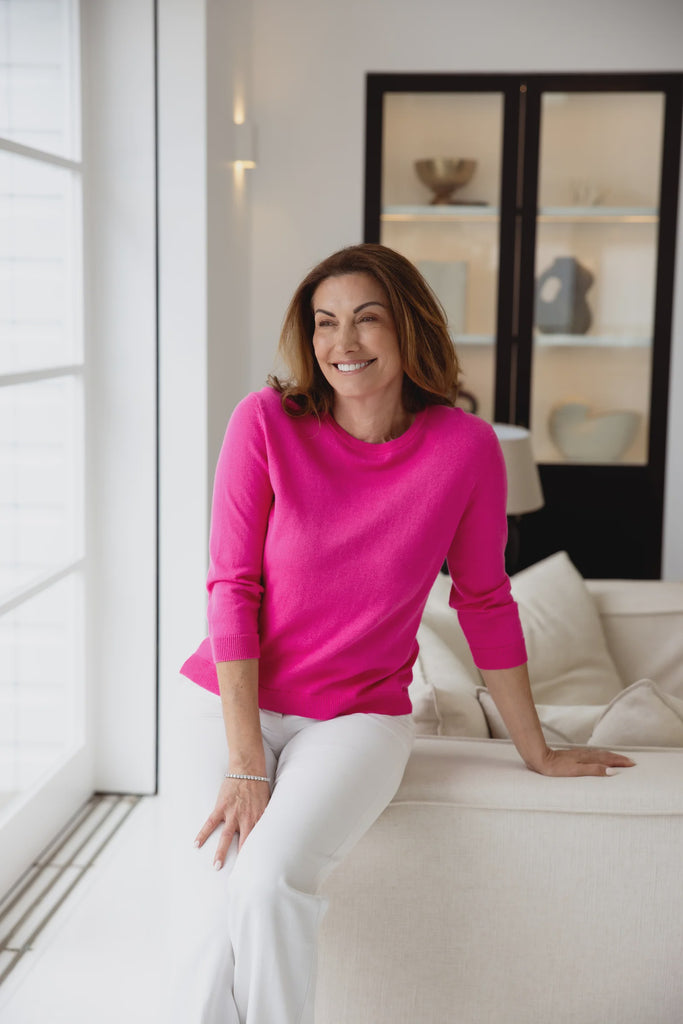 Woman in a bright pink sweater sitting on a couch in a modern living room. Alashan Avery 3/4 Sleeve top in Malibu pink