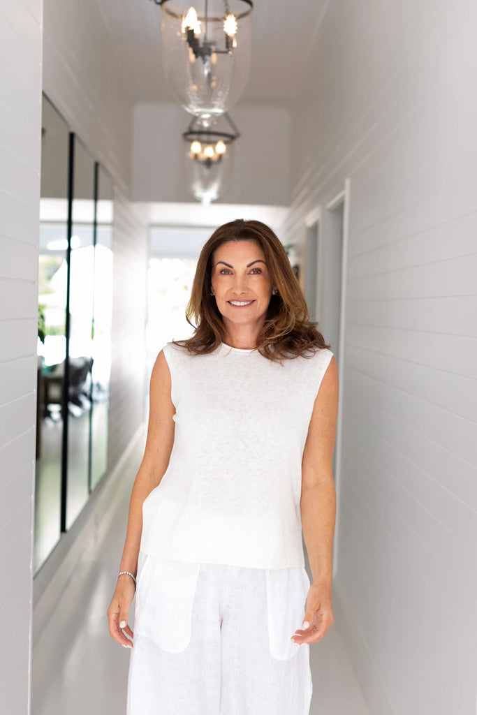 Woman in a white sleeveless top and pants standing in a hallway with a chandelier above. Alashan Crest Linen Tank in French Vanilla.