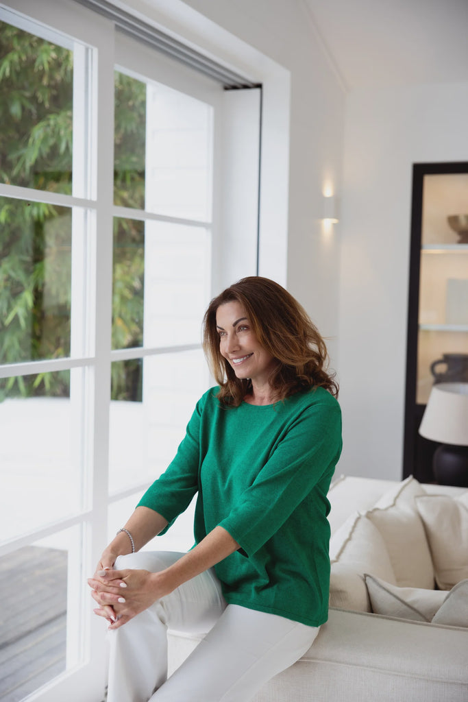 Woman in a green sweater sitting on a couch by a large window with a view of greenery. Alashan Everyday Tee in Emerald Green.