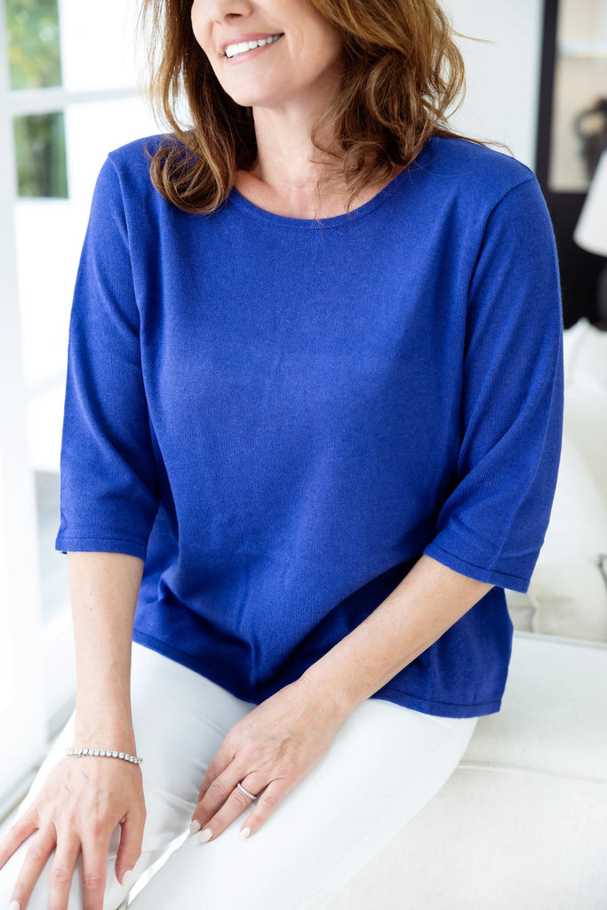 Woman in a blue top and white pants sitting on a couch in a modern living room. Alashan Everyday Tee in Royal.