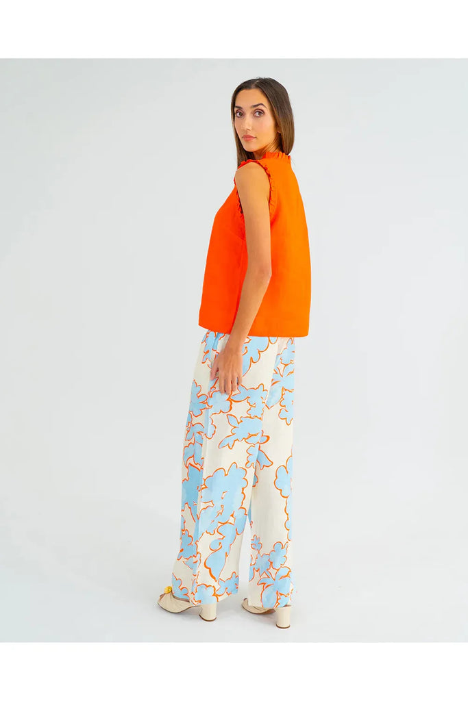 Woman wearing an orange sleeveless top with ruffled details on a plain background. Arlington Milne Lucia Top in Tangerine worn with the Bianca Trouser in Summertime Floral.