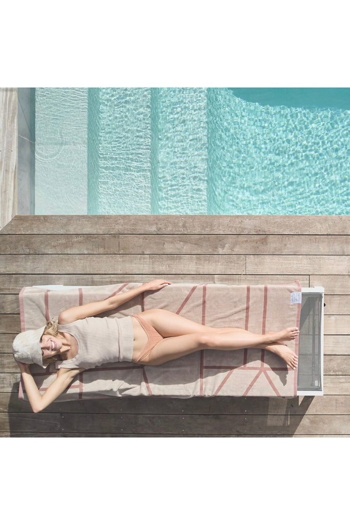& Sunday Journey Organic Beach/Pool Towel Dune Model laying on towel on a lounger beside Pool . Towel is reverse side up showing Cream with Dune Blush Lines 