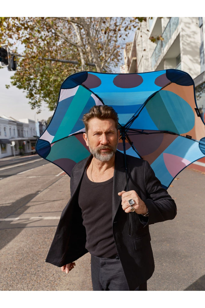 Blunt Classic Umbrella Limited Edition collab with Daimon Downey. Daimon Downey walking along a city street holding the open colourful umbrella over his left shoulder so the framework of the umbrella is visible.