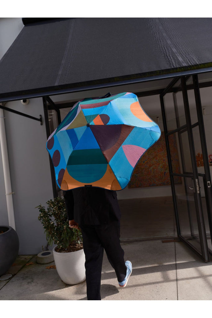 A person walking into a building entrance holding the colourful geometric-patterned Limited Edition Classic Blunt Umbrella collab with Daimon Downey. The image shows the colourful canopy of the umbrella over the person's shoulders. 
