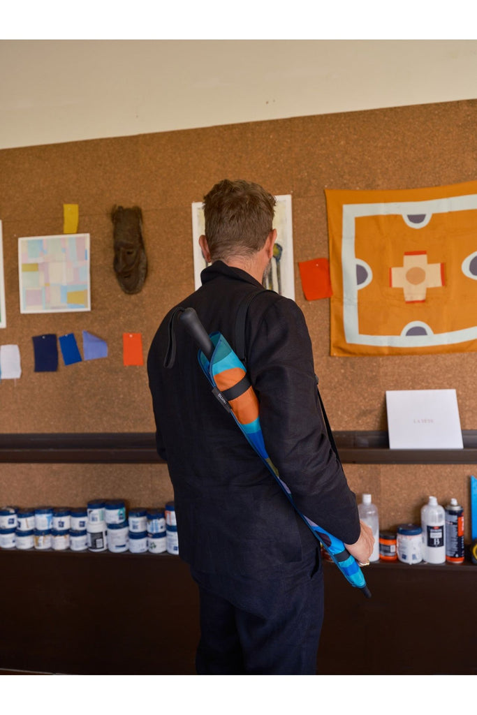 A man stands in front of a cork bulletin board with various items pinned to it, holding the colourful, limited edition Blunt and Daimon Downey Classic umbrella in its matching carry sleeve.