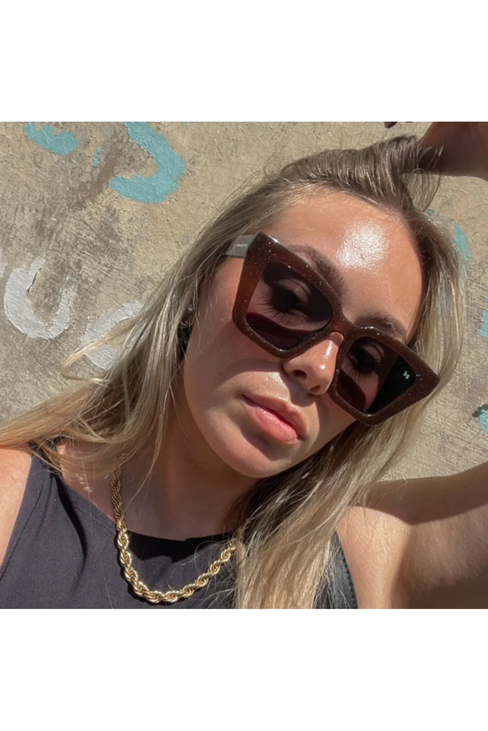 Woman wearing sunglasses and a gold necklace, lying on sand with a patterned towel. Bored George Piper Sunglasses in Shimmer.