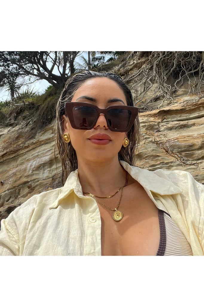 Woman wearing sunglasses and a light-colored shirt against a rocky background.  Bored George Piper Sunglasses in Shimmer.