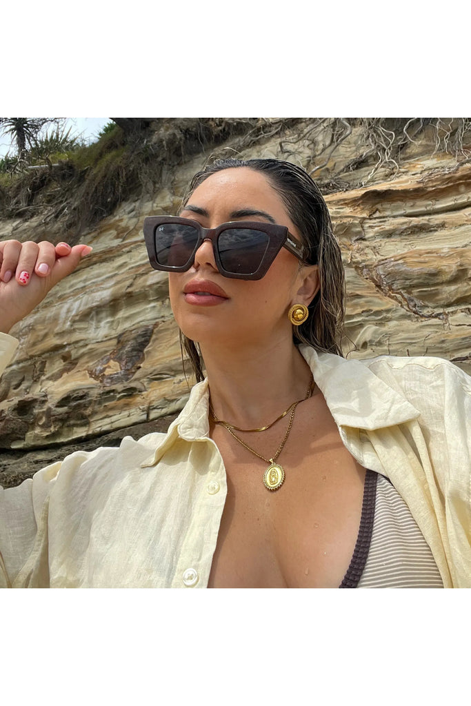Woman wearing sunglasses and a light-colored shirt with a rocky background. Bored George Piper Sunglasses in Shimmer.