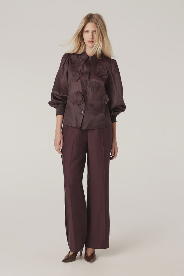 Woman wearing a brown blouse with floral embellishments and matching pants on a plain background. Cable Goldie Pant and Mirage Blouse in Raisin.