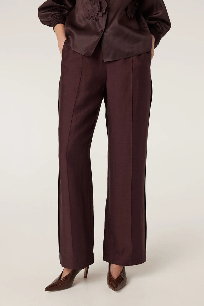 Woman wearing a brown blouse with floral embellishments and matching pants on a plain background. Cable Goldie Pant and Mirage Blouse in Raisin.