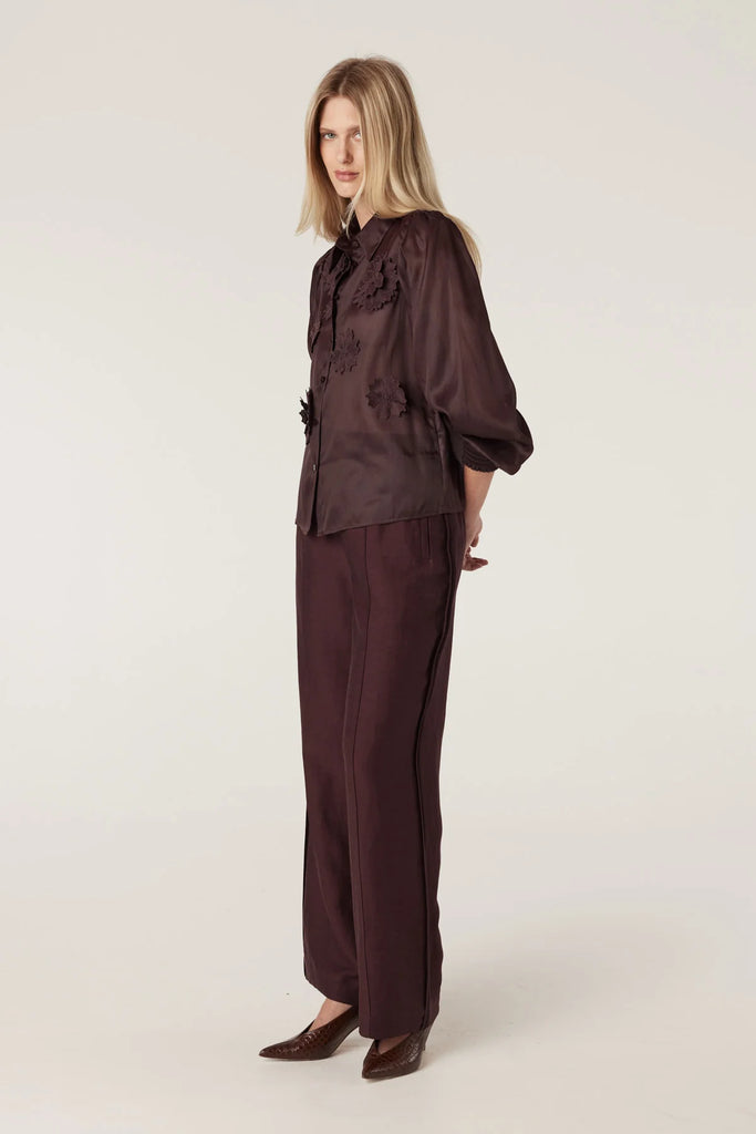 Woman wearing a brown blouse with floral embellishments and matching pants on a plain background. Cable Goldie Pant and Mirage Blouse in Raisin.