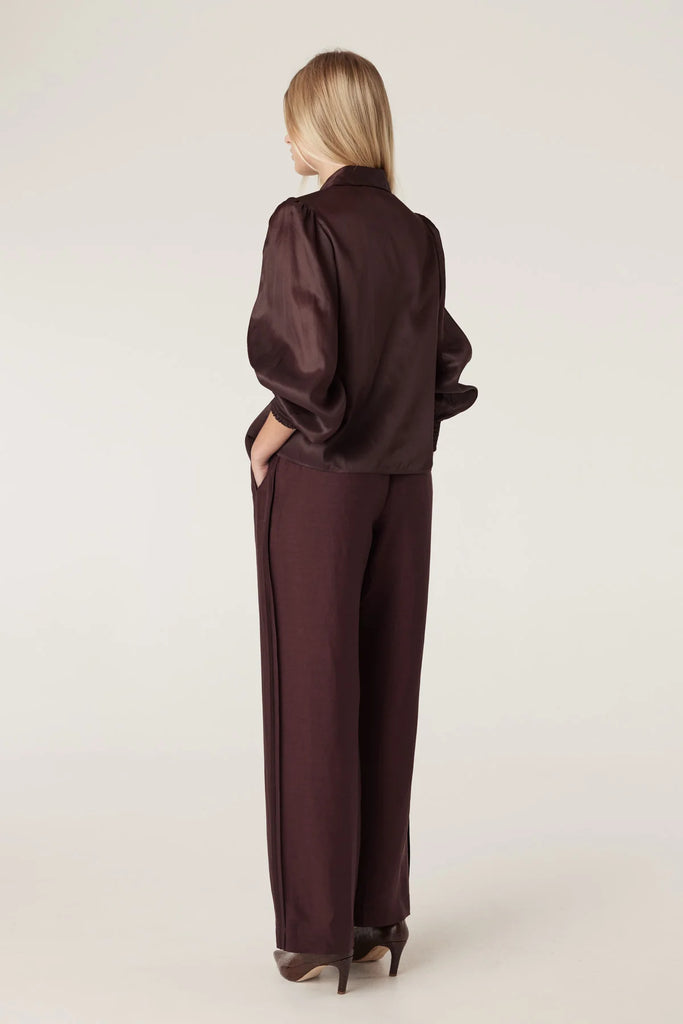 Woman wearing a brown blouse with floral embellishments and matching pants on a plain background. Cable Goldie Pant and Mirage Blouse in Raisin.