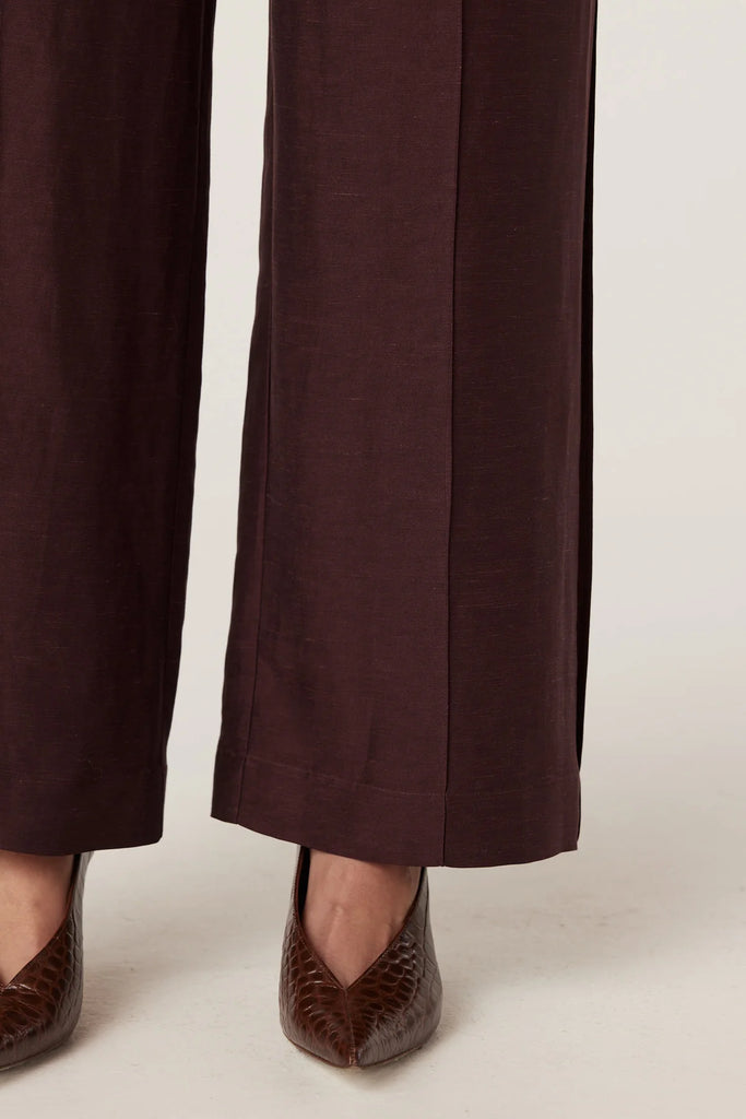 Woman wearing a brown blouse with floral embellishments and matching pants on a plain background. Cable Goldie Pant and Mirage Blouse in Raisin.