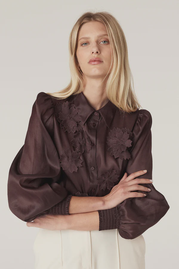 Woman wearing a dark brown blouse with floral details on a plain background. Cable Mirage Blouse in raisin.