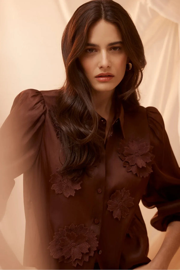 Woman wearing a dark brown blouse with floral details on a plain background. Cable Mirage Blouse in raisin.