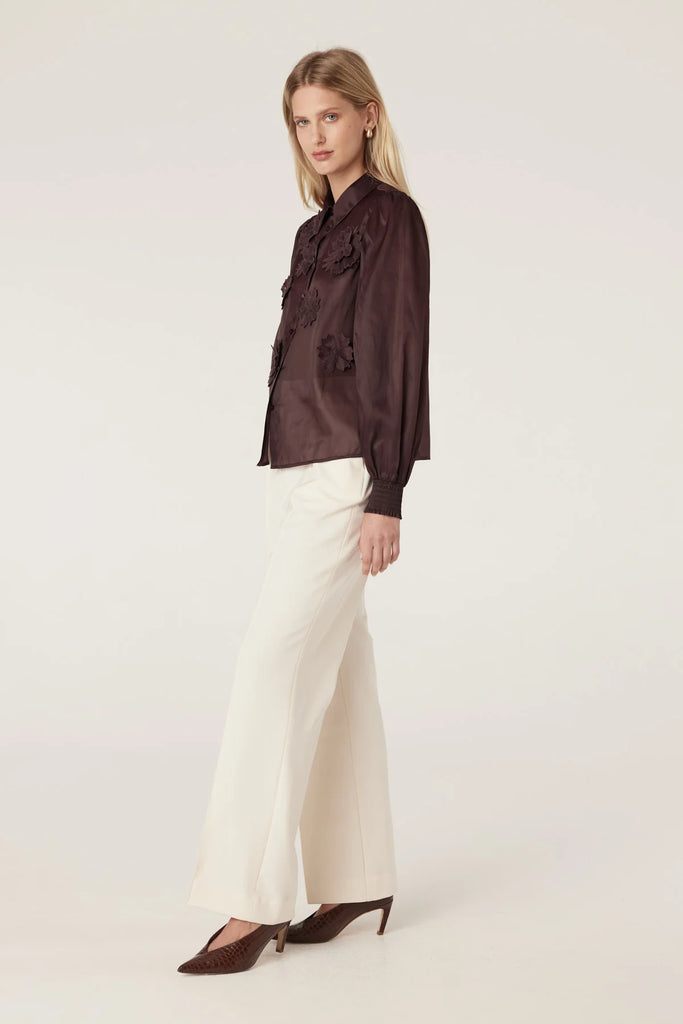 Woman wearing a dark brown blouse with floral details on a plain background. Cable Mirage Blouse in raisin.