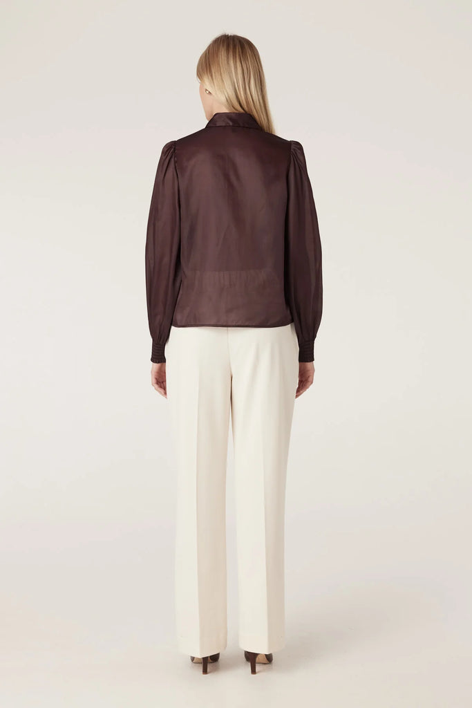 Woman wearing a dark brown blouse with floral details on a plain background. Cable Mirage Blouse in raisin.