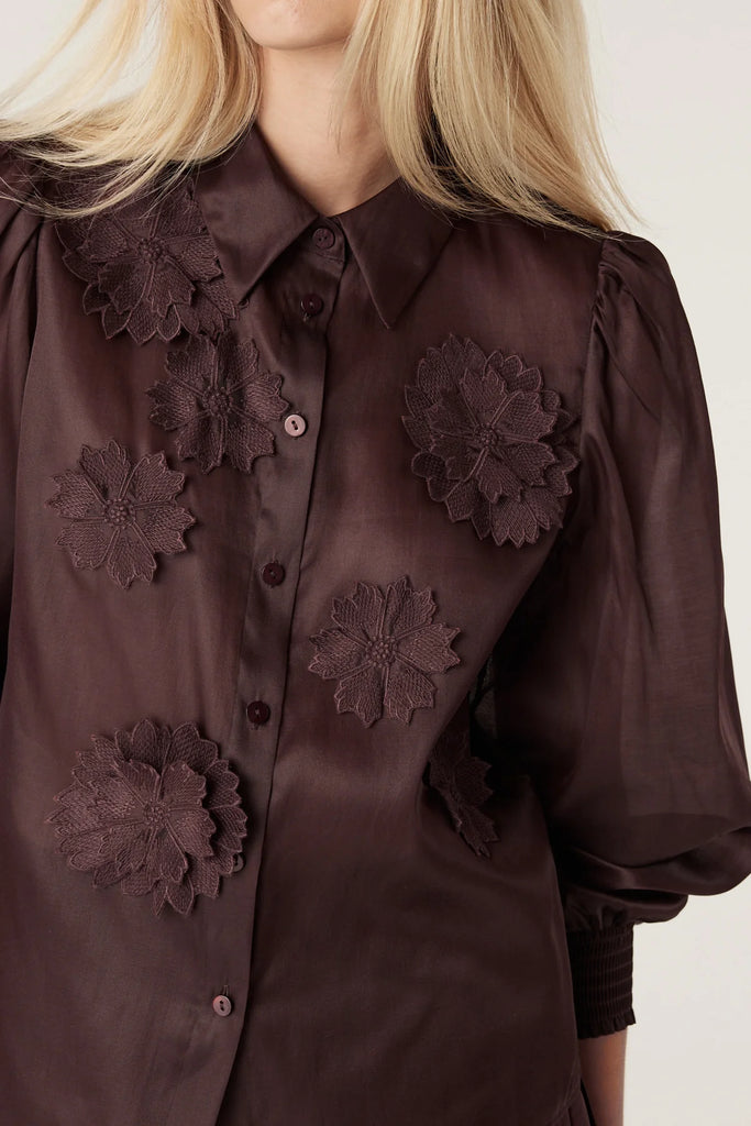 Woman wearing a dark brown blouse with floral details on a plain background. Cable Mirage Blouse in raisin.