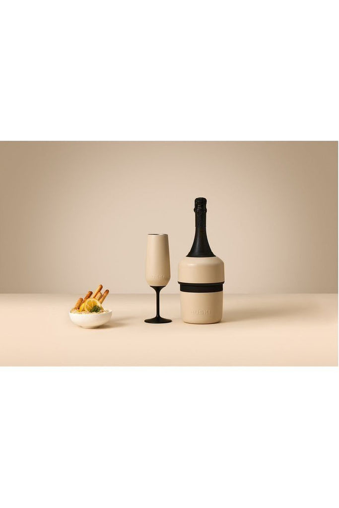 The exclusive Huski Champagne Cooler in sand beige beautifully complements the sand beige Huski Champagne Tumbler with its elegant black stem, all showcased against a delightful beige background filled with carefully arranged food.