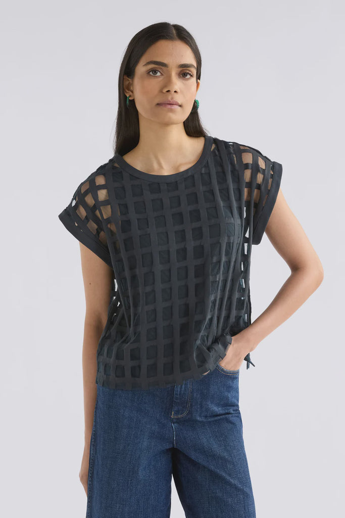Woman wearing a dark blue patterned top and blue jeans on a light gray background. Elk Grid Burnout top in Space Blue.