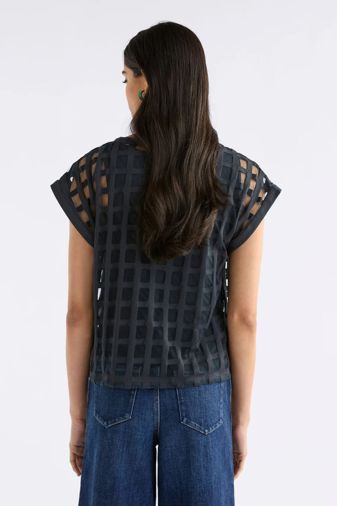 Woman wearing a dark blue patterned top and blue jeans on a light gray background. Elk Grid Burnout top in Space Blue.