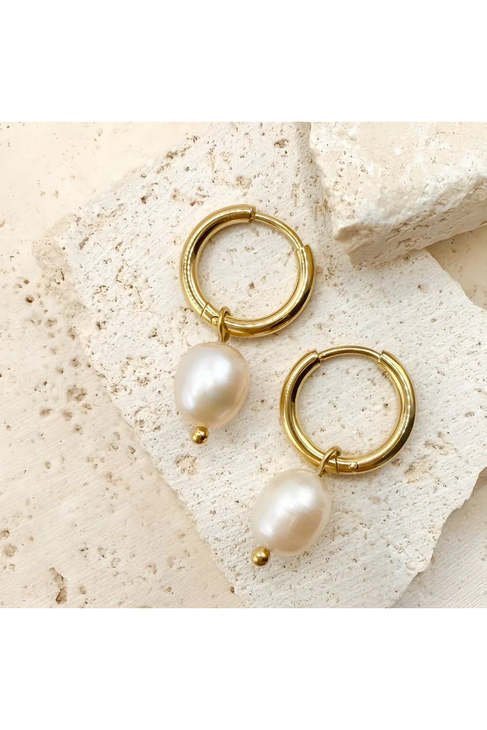 Gold hoop earrings with pearl drops on a textured beige surface. After Dark Pearl Hoop Earrings in gold by Ever.