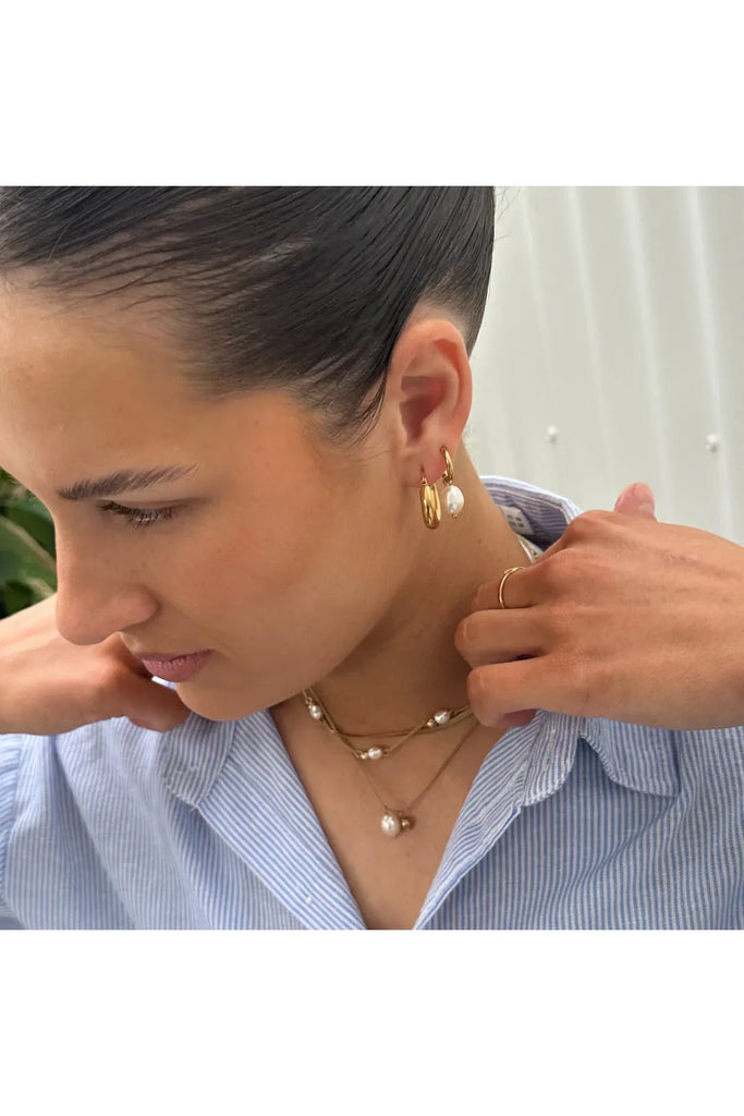 Woman wearing gold hoop earrings and pearl necklaces, adjusting her collar. After Dark Pearl Hoop Earrings in gold by Ever.