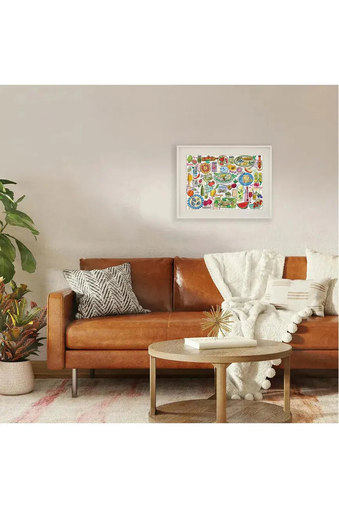 Piece House 500-piece jigsaw puzzle titled 'Feast & Fizz' in collaboration with Jo Tronc. Once completed, the framed puzzle is displayed in a living room above a brown leather sofa.