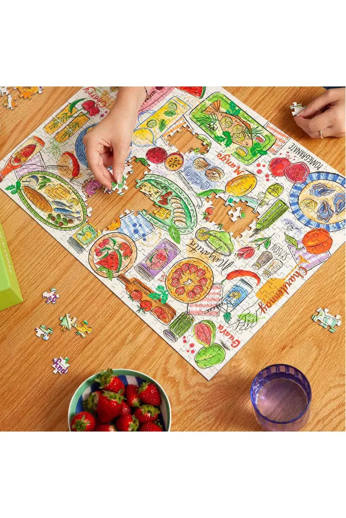 Piece House 500-piece jigsaw puzzle box titled 'Feast & Fizz' in collaboration with Jo Tronc. A person assembling this colourful puzzle on a wooden table with a bowl of strawberries and a glass.
