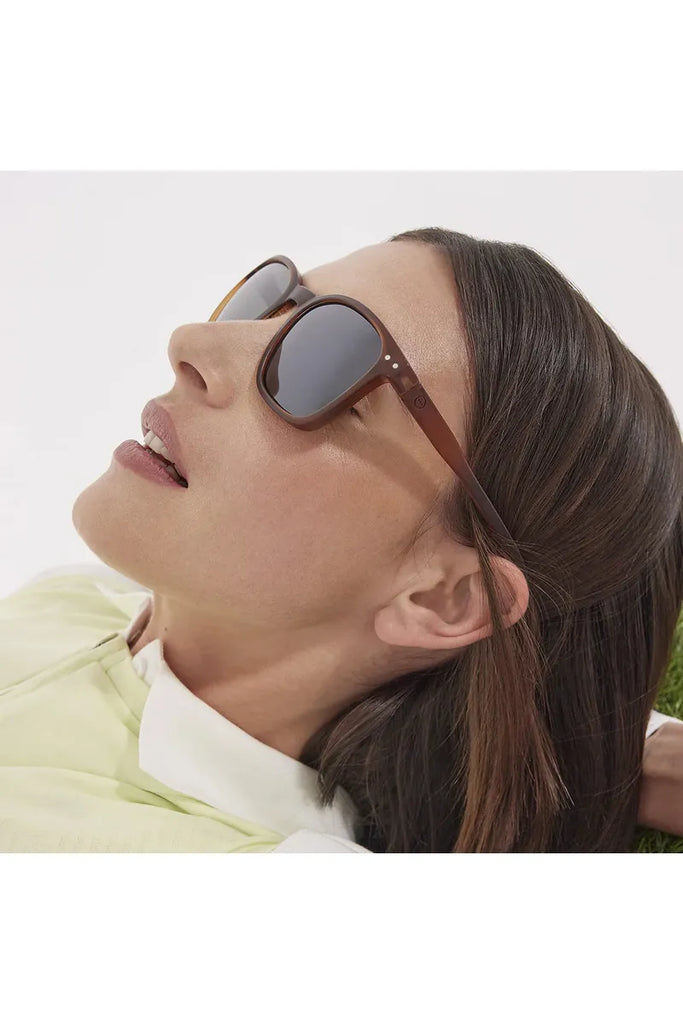 A woman wearing Izipizi hazel brown polarised sunglasses lies on the grass with a white background.