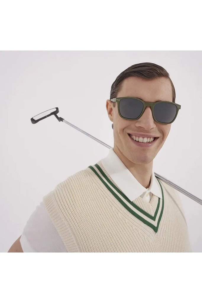 A man wearing Izipizi polarised khaki green sunglasses and a cream sweater vest with a white shirt, holding a golf putter.