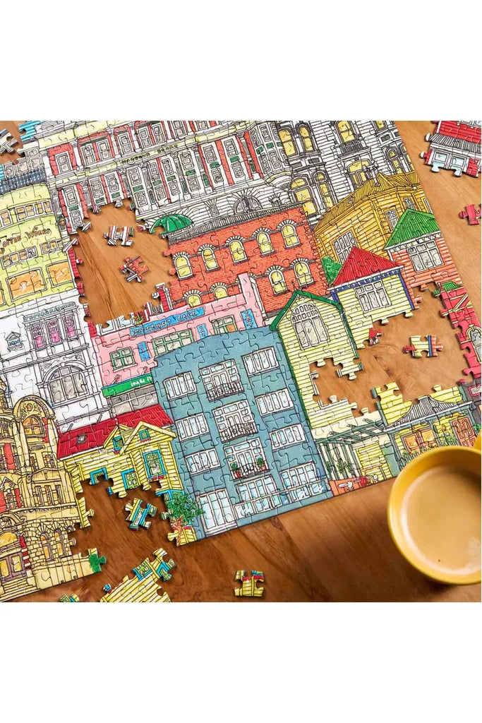 Piece House is collaborating with Stacey Fraser Allen on a puzzle titled "Neighbourhood Puzzle." The image depicts a puzzle of the Wellington cityscape on a wooden table with a cup of coffee.