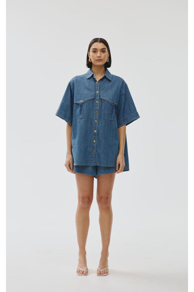 Woman wearing a blue denim outfit on a white background. Kinney Juniper Shorts with the matching Bella Shirt in a light denim fabric.