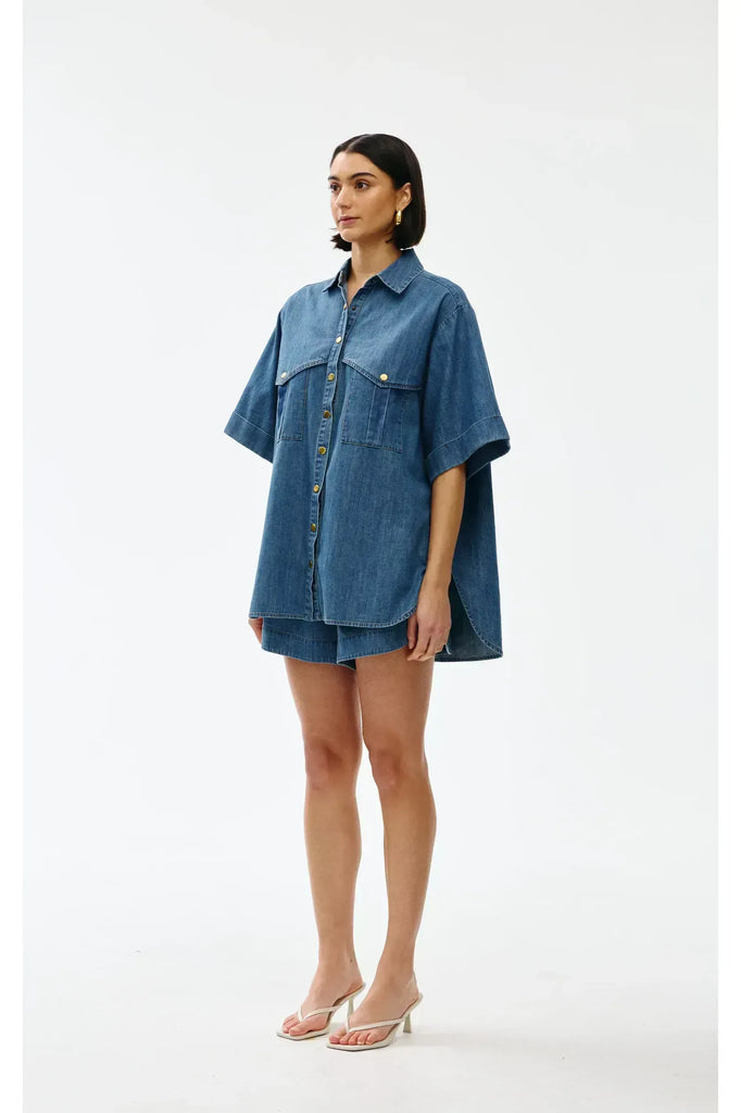 Woman wearing a blue denim outfit on a white background. Kinney Juniper Shorts with the matching Bella Shirt in a light denim fabric.