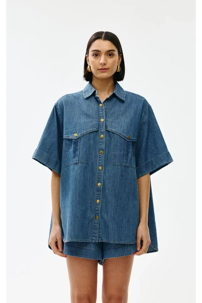 Woman wearing a blue denim outfit on a white background. Kinney Juniper Shorts with the matching Bella Shirt in a light denim fabric.