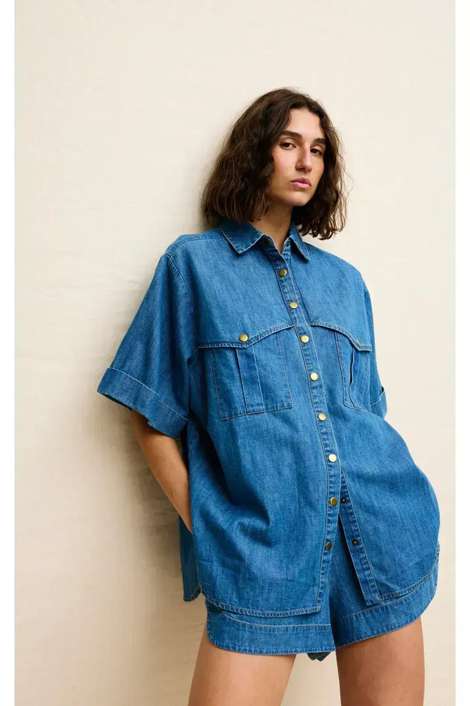 Woman wearing a blue denim outfit on a white background. Kinney Juniper Shorts with the matching Bella Shirt in a light denim fabric.