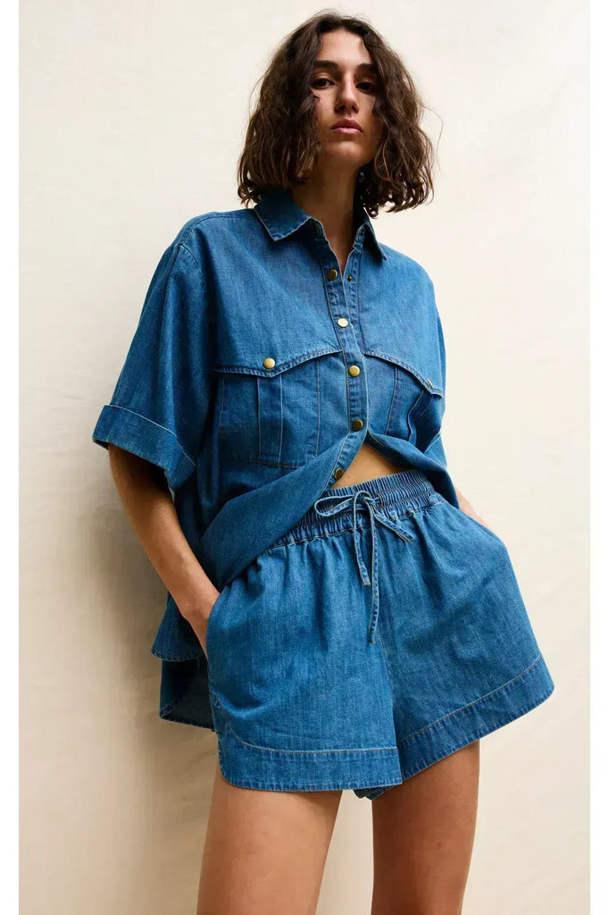 Woman wearing a blue denim outfit on a white background. Kinney Juniper Shorts with the matching Bella Shirt in a light denim fabric.